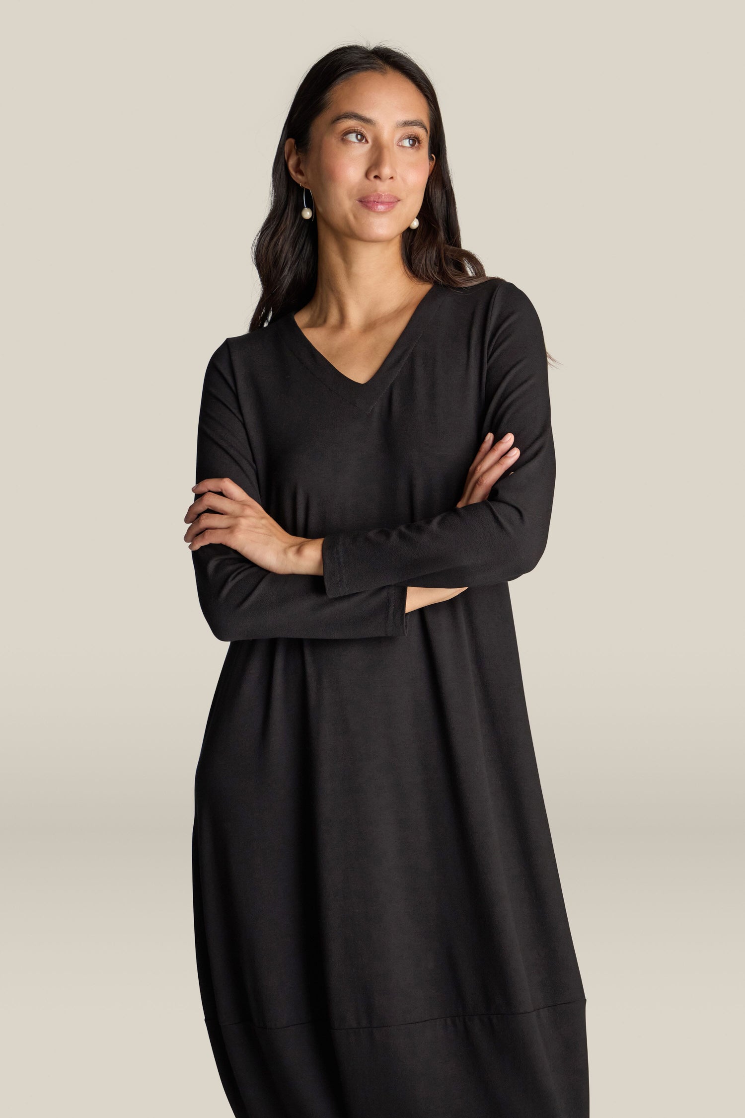 A woman with long dark hair stands with arms crossed, wearing the Fluid Crepe V Neck Bubble Dress. The universally flattering silhouette of this simple black dress, featuring long sleeves and a V-neck, accentuates her form as she looks slightly to the side.