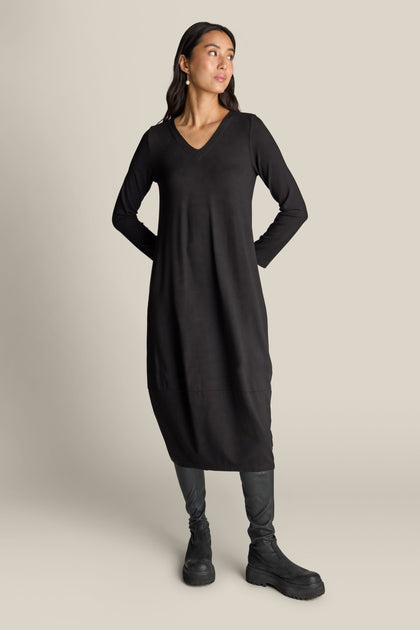 A woman stands wearing a Fluid Crepe V Neck Bubble Dress in long-sleeve black, paired with black boots. She looks away from the camera with her hands behind her back, showcasing its universally flattering silhouette. Her long dark hair flows against the neutral background.