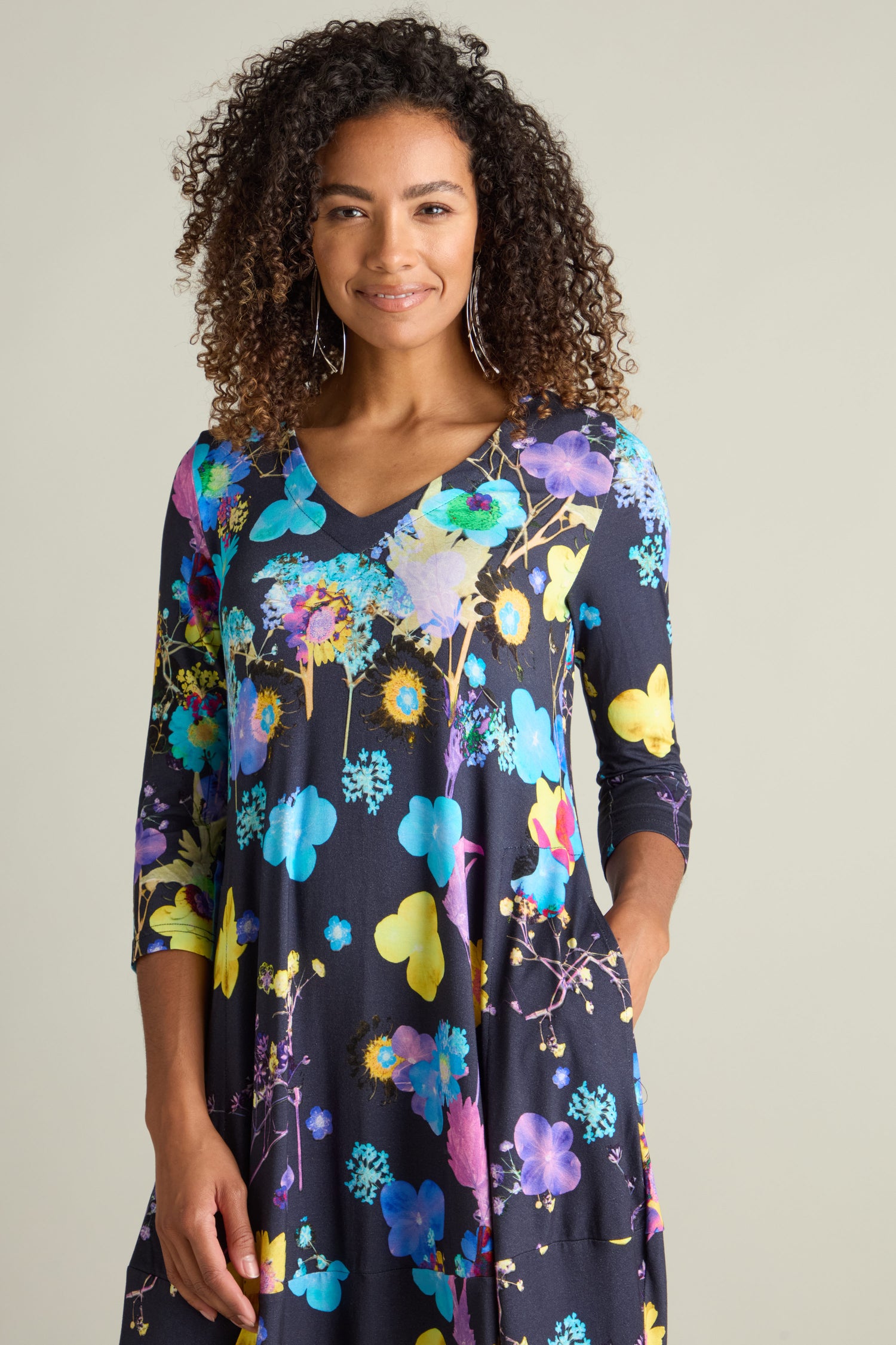 A person with curly hair wearing the Blue Bouquet Jersey Dress, which features a colorful botanical print, stands against a plain background.