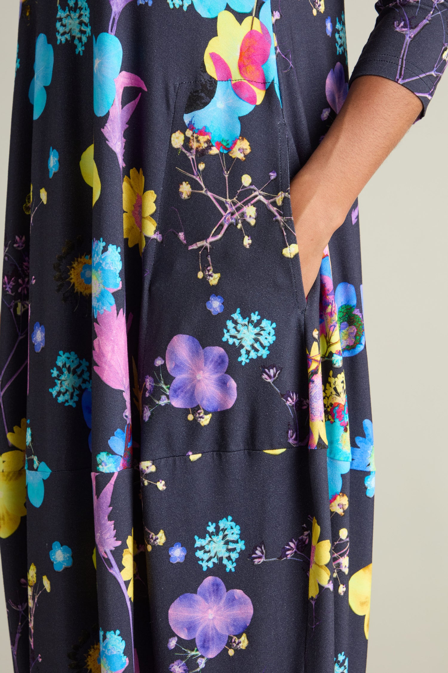 Close-up of a person's hand in their dress pocket. The Blue Bouquet Jersey Dress showcases a black fabric adorned with a vibrant botanical print, crafted from soft stretch jersey.