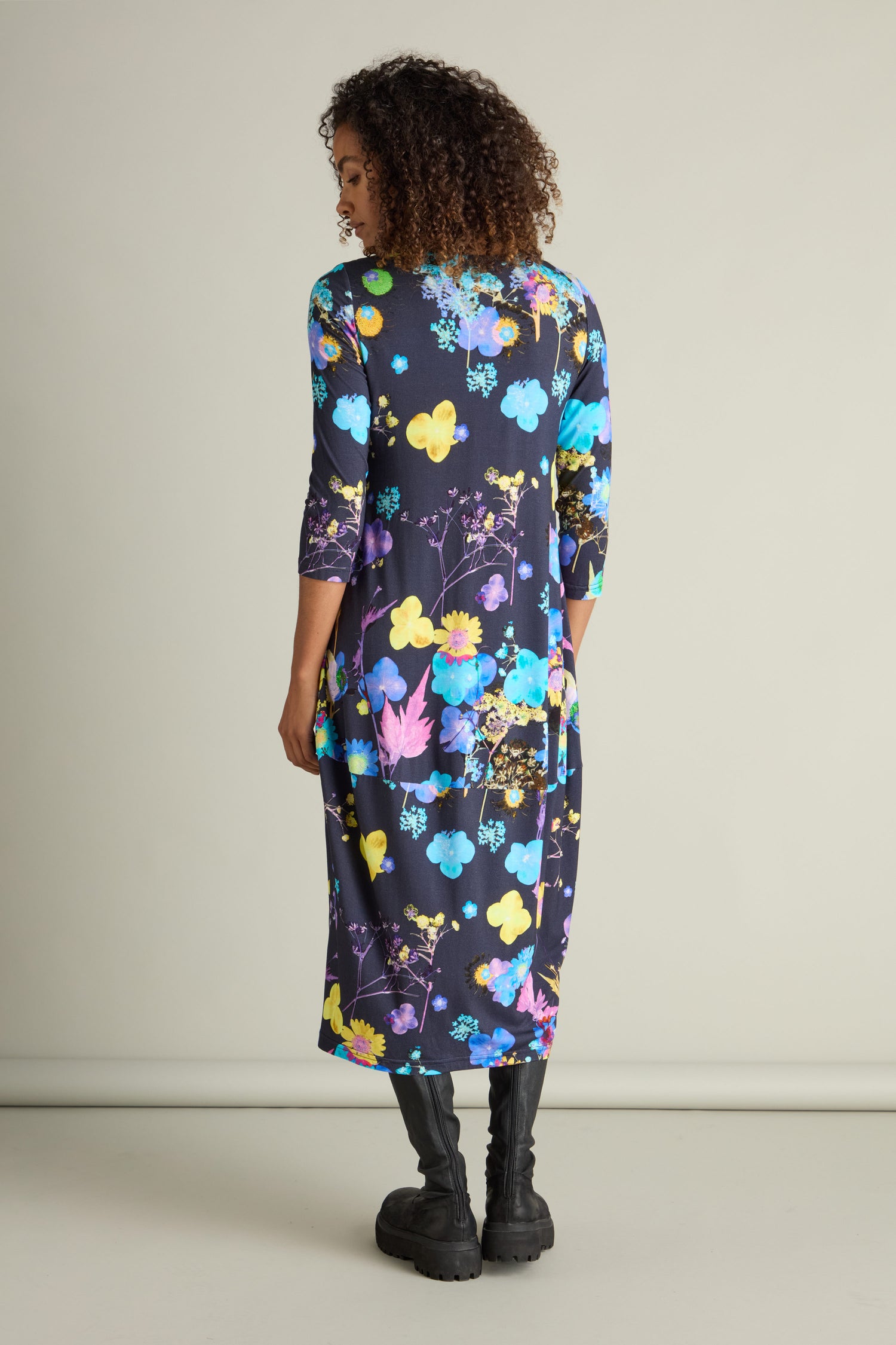 A person stands facing away, wearing the Blue Bouquet Jersey Dress adorned with a delicate botanical print and paired with black boots, set against a plain background.