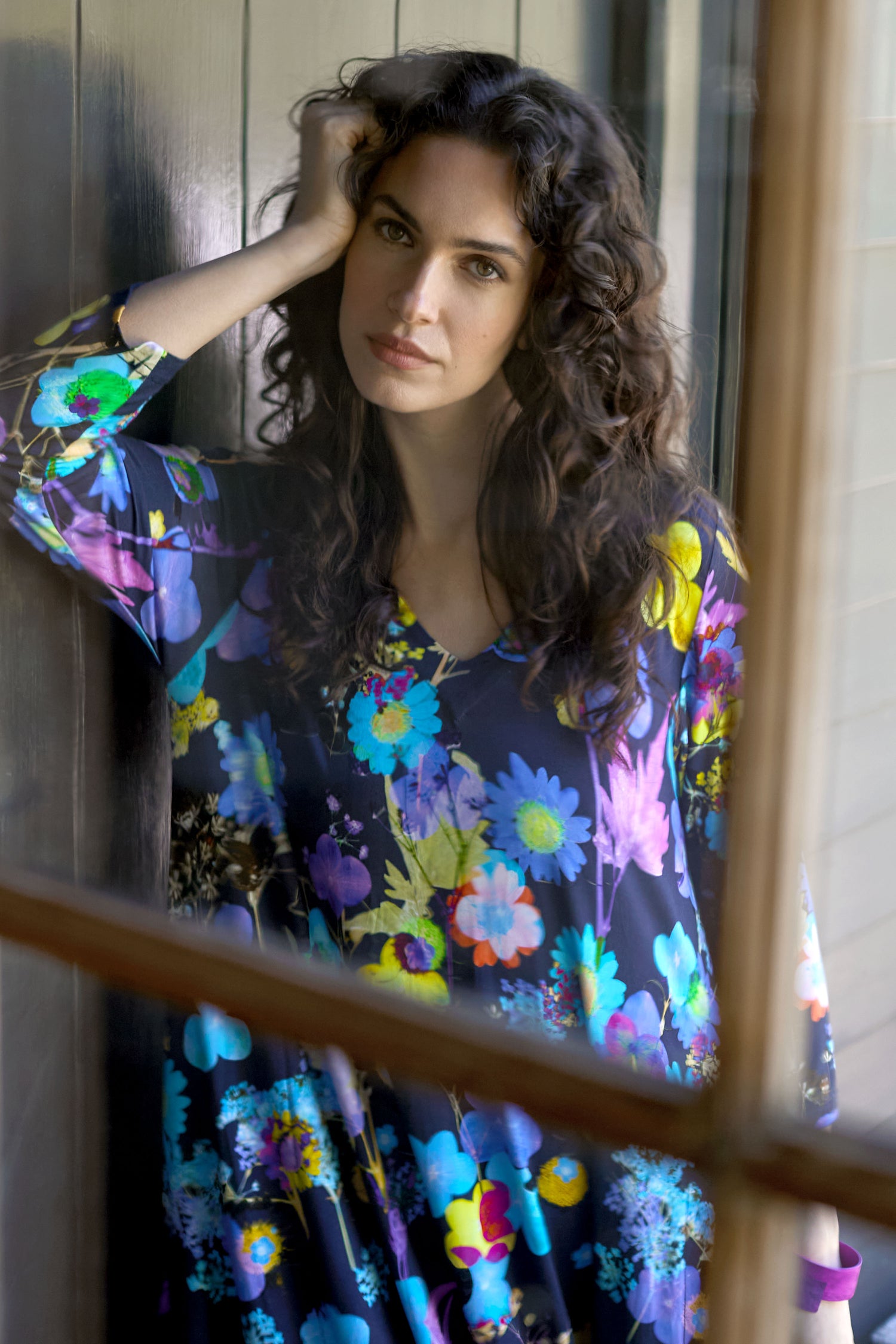 A woman with long, curly hair leans against a wall seen through a window, wearing the Blue Bouquet Jersey Dress adorned with a vibrant botanical print.