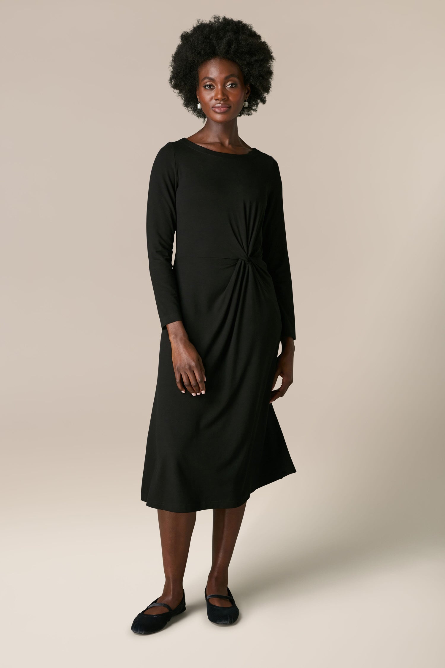 A woman with short curly hair is wearing a Fluid Crepe Knot Dress featuring full-length sleeves as she stands against a beige background.