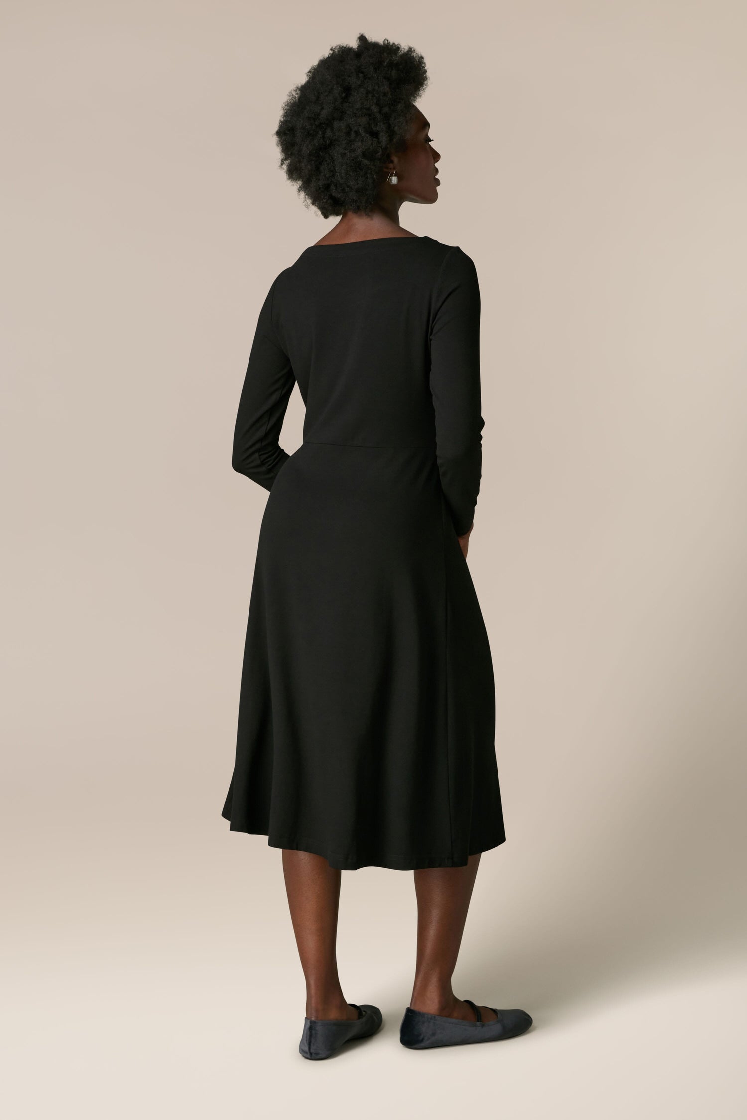 A person with short curly hair is standing and facing away, wearing a Fluid Crepe Knot Dress with full-length sleeves and dark shoes against a plain background.