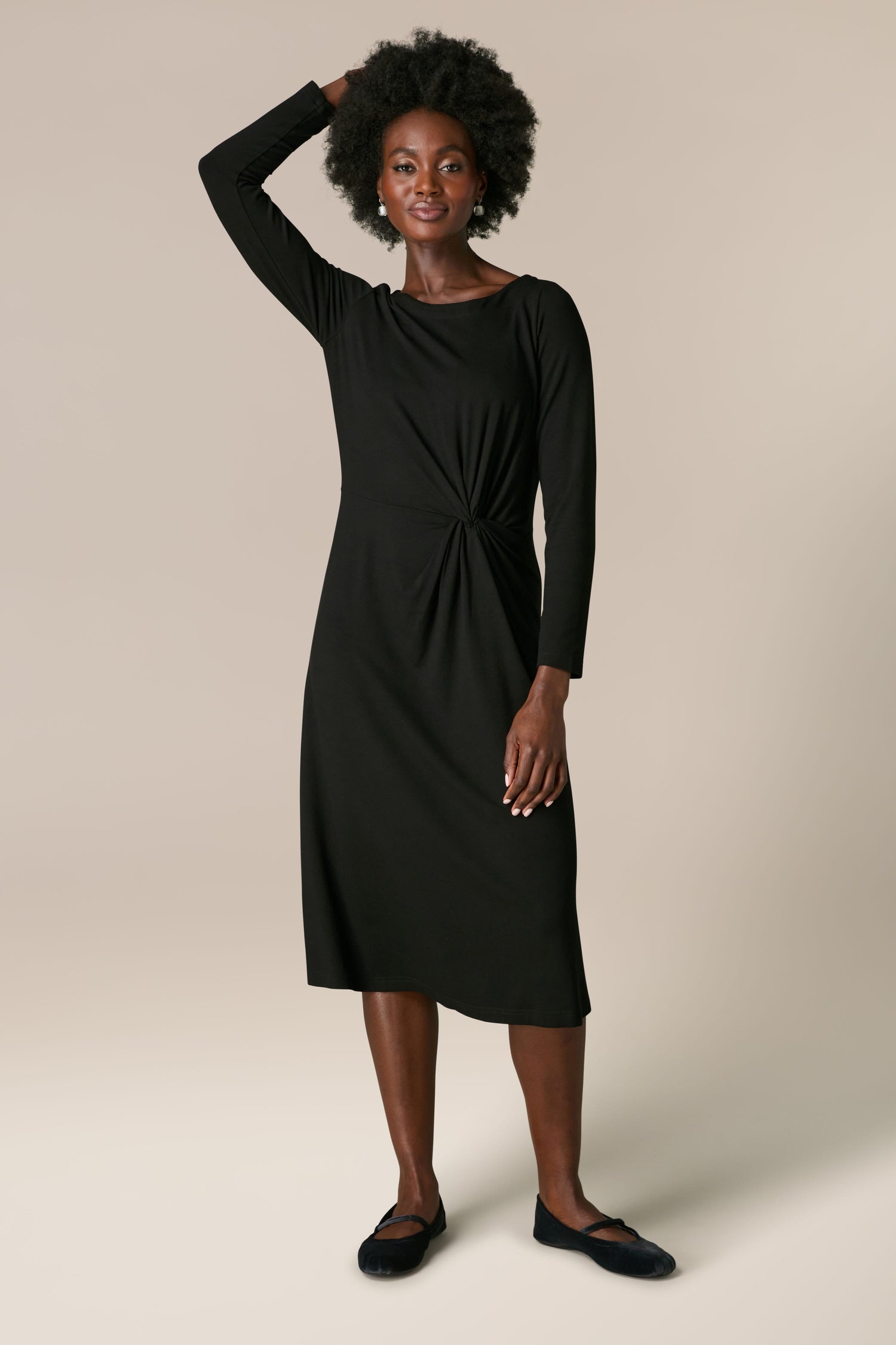 A woman stands against a neutral background, wearing a sophisticated Fluid Crepe Knot Dress with full-length sleeves and matching shoes. She has one hand resting on her head.