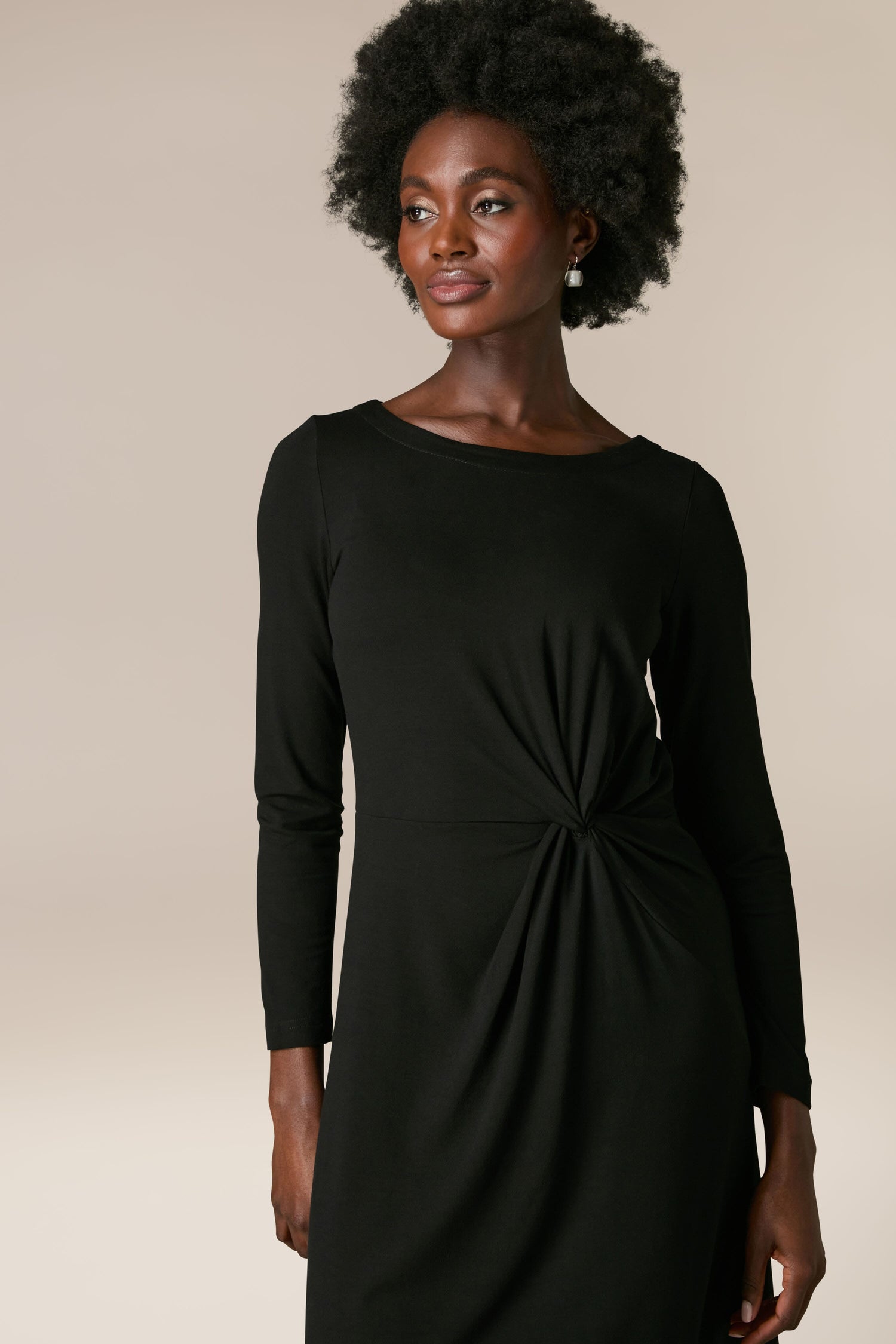 A woman wearing a Fluid Crepe Knot Dress with full-length sleeves and a knot detail at the waist poses gracefully against a plain beige background.