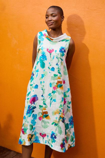 A person stands against an orange wall, wearing a sleeveless Watercolour Foxglove Linen Dress. Their hands are resting behind their back.