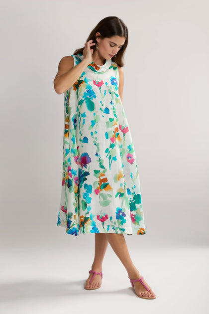 A woman wearing a sleeveless Watercolour Foxglove Linen Dress with colorful patterns stands against a plain background, looking down and adjusting her hair with her right hand. Her pink sandals add an elegant charm to the classic A-line sheath dress.