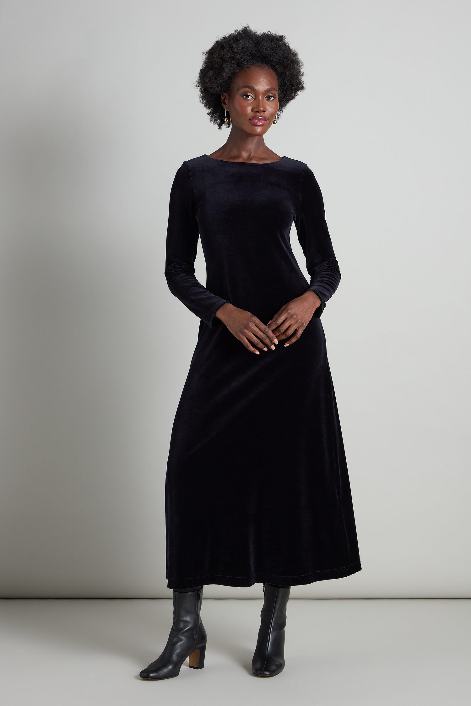 A person exudes understated glamour in a Velvet Jersey Maxi A-Line Dress, paired with black boots, against a plain light gray background.