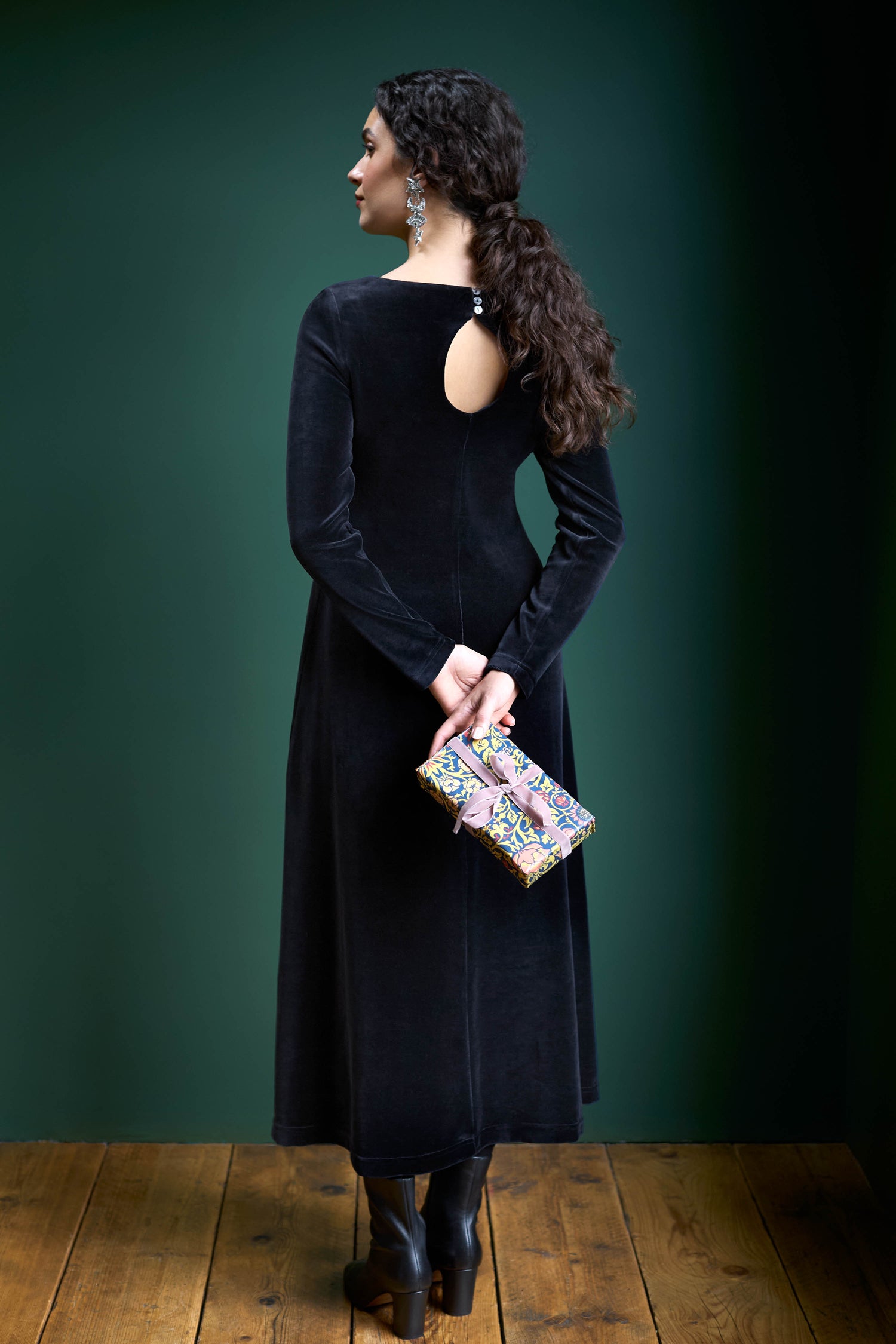 The woman stands with her back to the camera, embodying understated glamour in a long-sleeved Velvet Jersey Maxi A-Line Dress. She clutches a colorful purse against the vibrant green background, effortlessly showcasing a fit-and-flare silhouette.