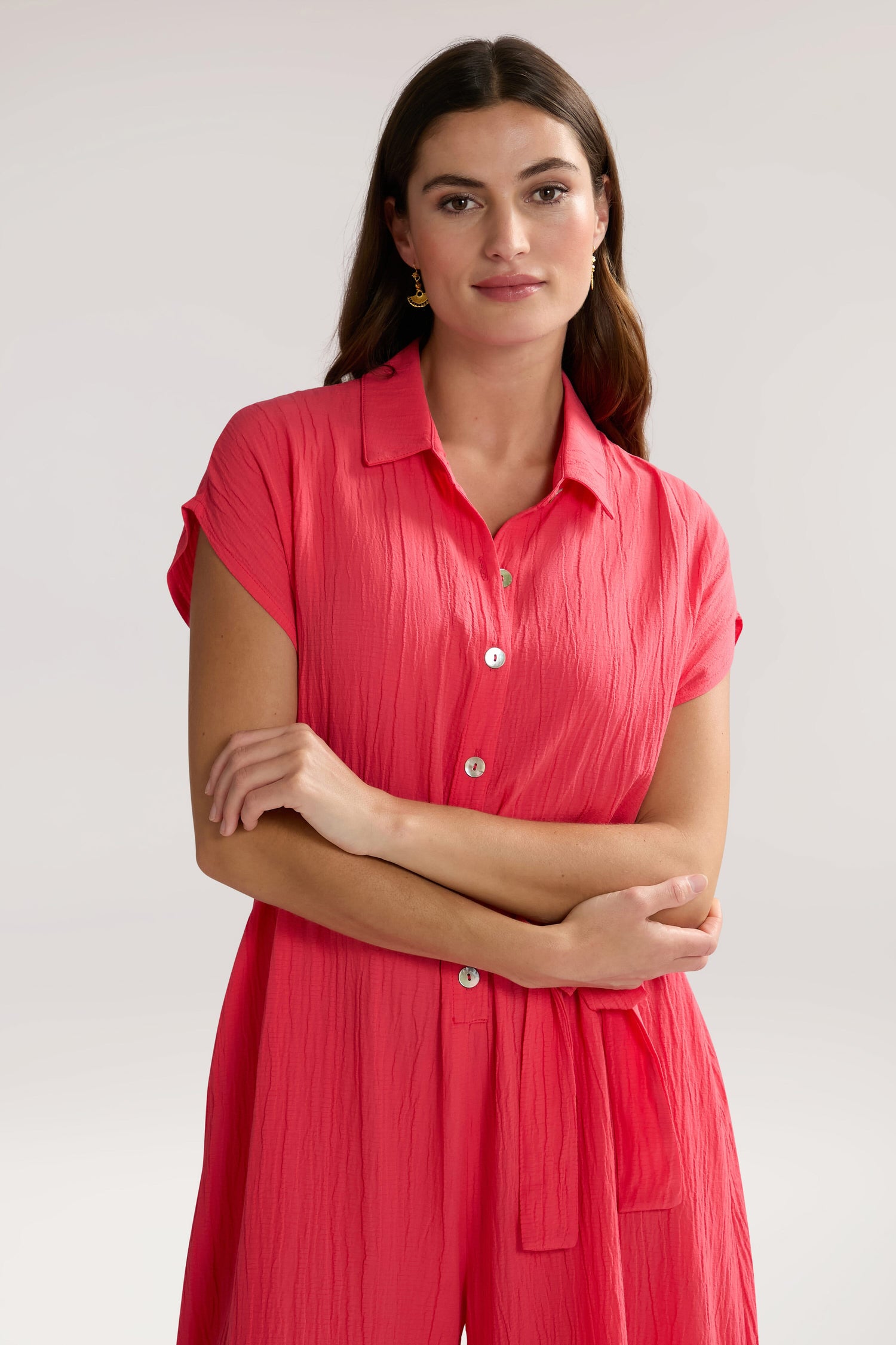 A woman with long hair wearing a bright pink, short-sleeve button-up Crinkle Viscose Jumpsuit stands with her arms crossed, looking at the camera.