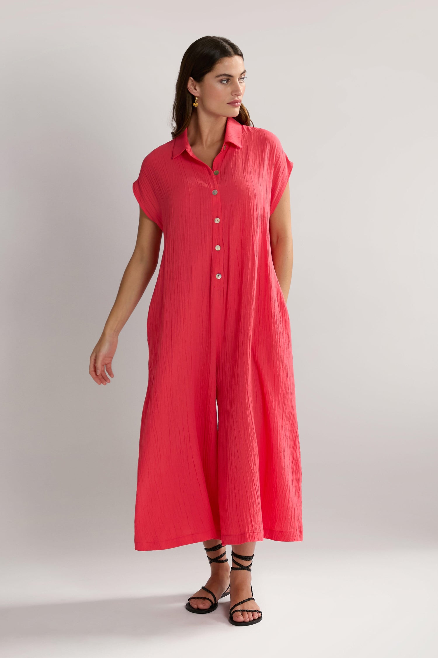 A person stands in a pink Crinkle Viscose Jumpsuit with short sleeves and button details, wearing black strappy sandals. The outfit is accented with an SPD5896-CSV tag, adding a touch of modernity to the classic look.