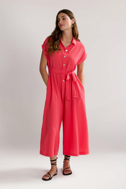 A woman stands against a plain background, wearing a vibrant pink Crinkle Viscose Jumpsuit with a belt, labeled SPD5896-CSV, along with black strappy sandals and hoop earrings. She gazes slightly to her left, with her hands in her pockets.