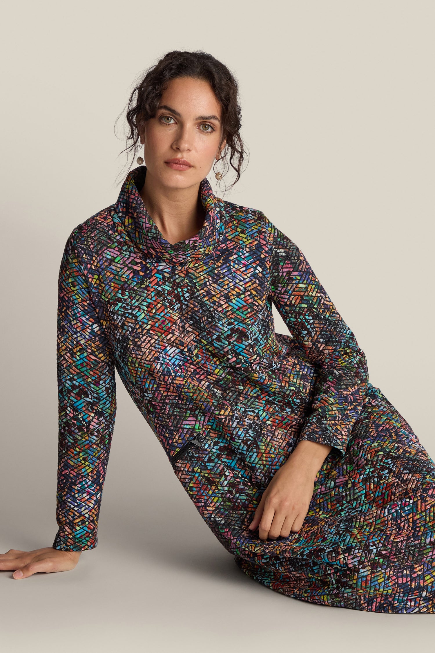 Woman sits against a plain backdrop wearing a Diamond Jacquard Jersey Cocoon Dress with a multicolored, geometric pattern.