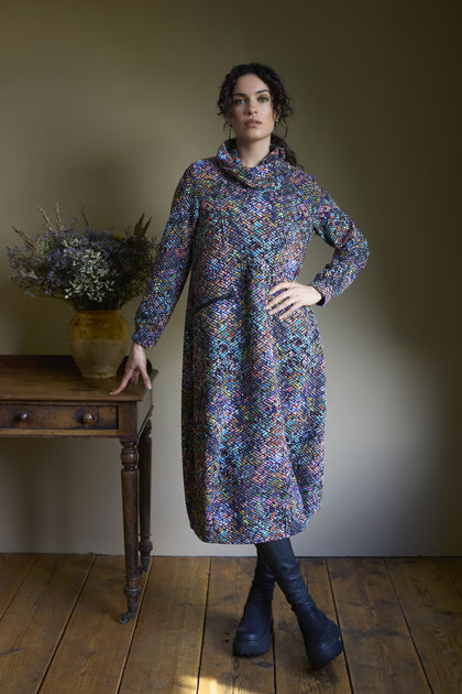 A woman in a vibrant Diamond Jacquard Jersey Cocoon Dress and black boots stands indoors next to a wooden table with a vase of flowers.