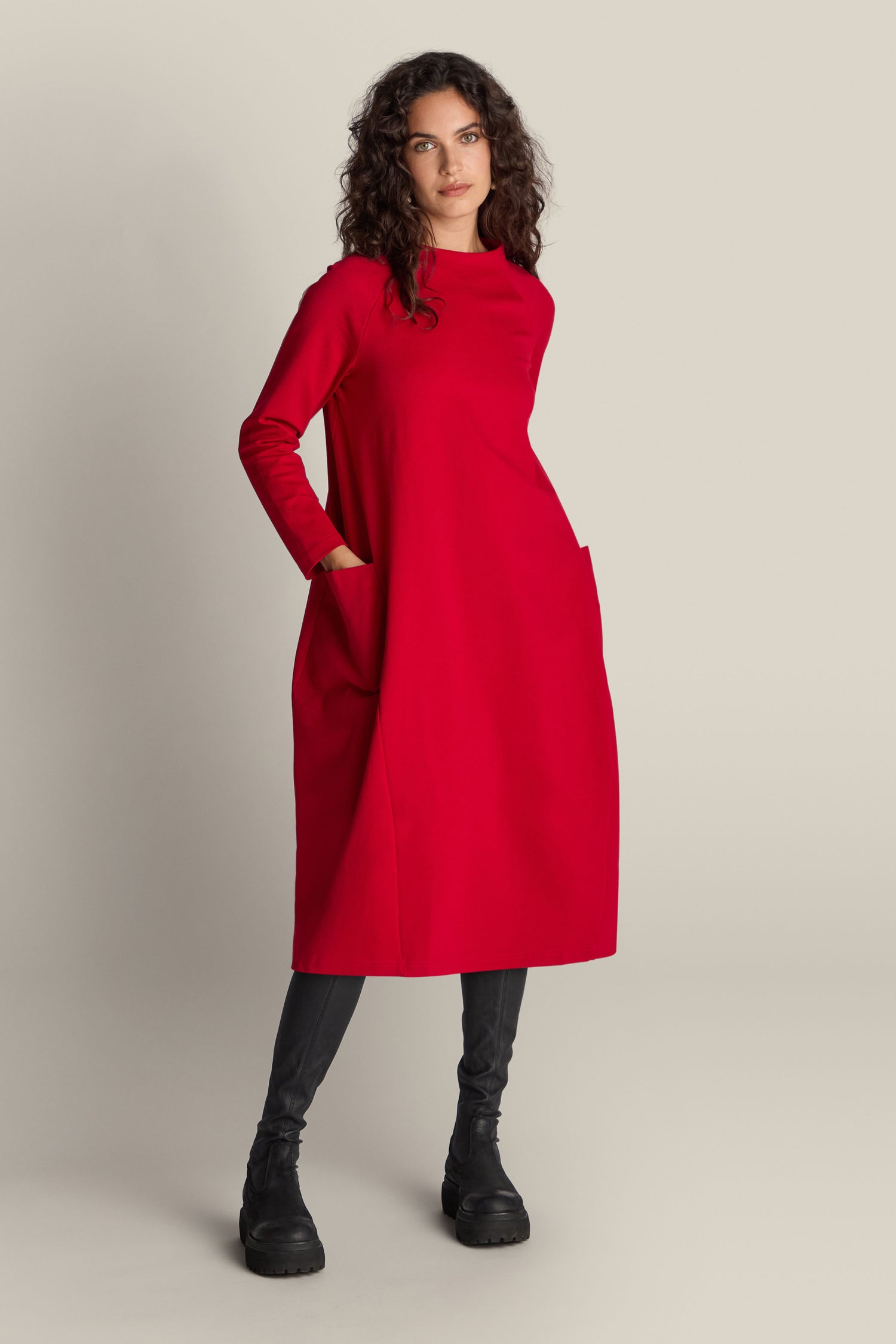 A woman stands wearing a long-sleeved red Ponte Pocket Dress, showcasing its versatile silhouette, paired with black leggings and black boots, against a plain background.