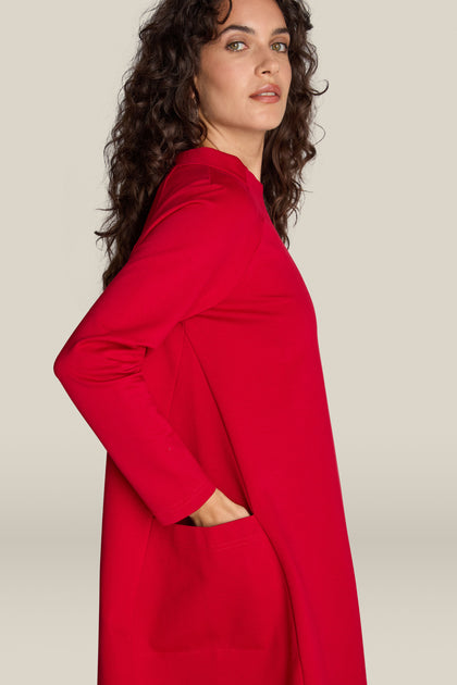 Profile view of a person with curly hair wearing the Ponte Pocket Dress, featuring long sleeves in a vibrant red hue. The individual has their left hand comfortably tucked in the dress pocket and gazes slightly upward. The dress boasts a versatile silhouette that flatters effortlessly, embracing a cocoon shape that adds to its charm.