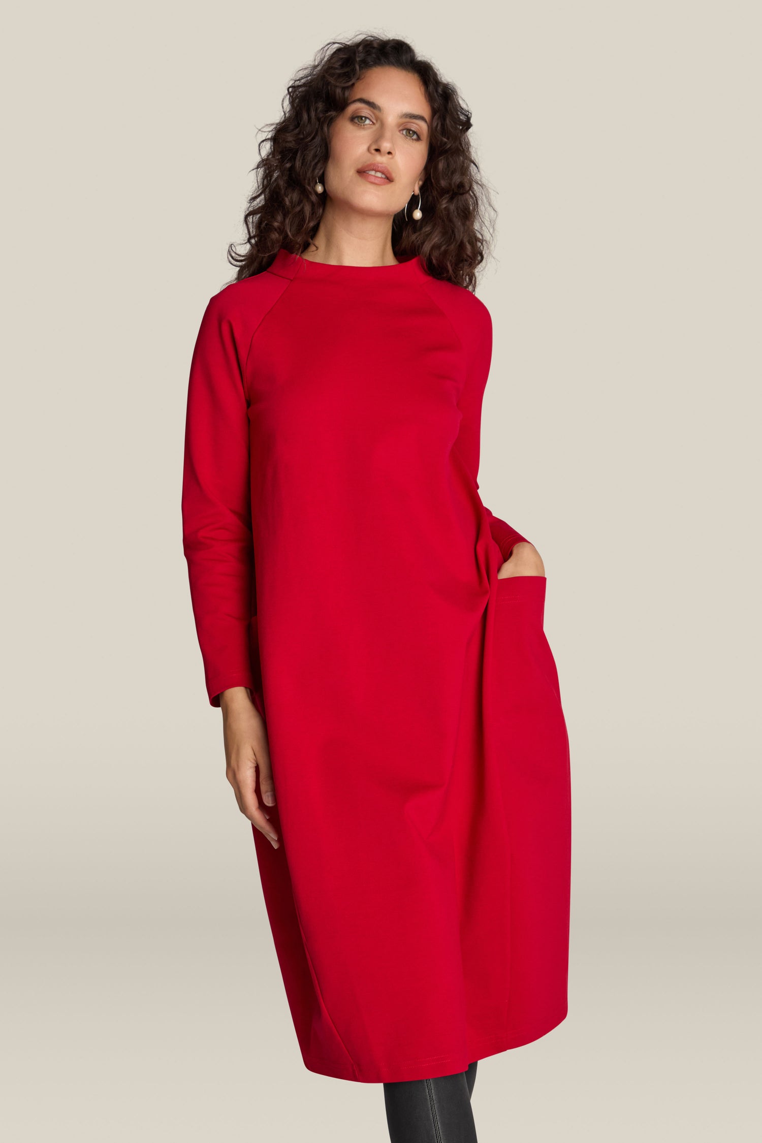 A person with curly hair is wearing the Ponte Pocket Dress, featuring long sleeves and a knee-length cut in red, with a versatile silhouette and has one hand in their pocket.