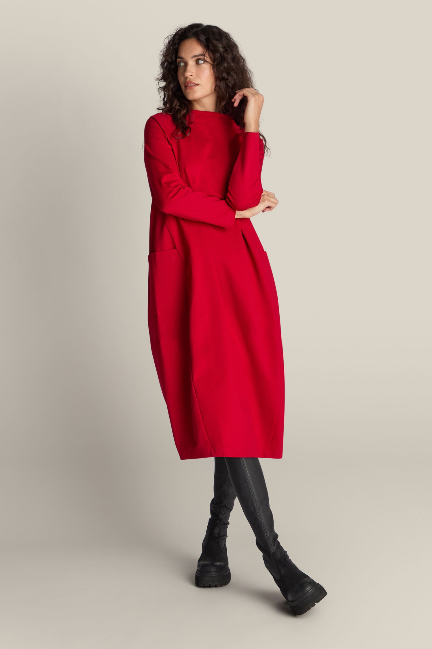 A woman with curly hair poses in a red, long-sleeve, knee-length Ponte Pocket Dress featuring pockets and black boots, against a plain, light-colored background. The versatile silhouette of the Ponte Pocket Dress beautifully flatters her figure.