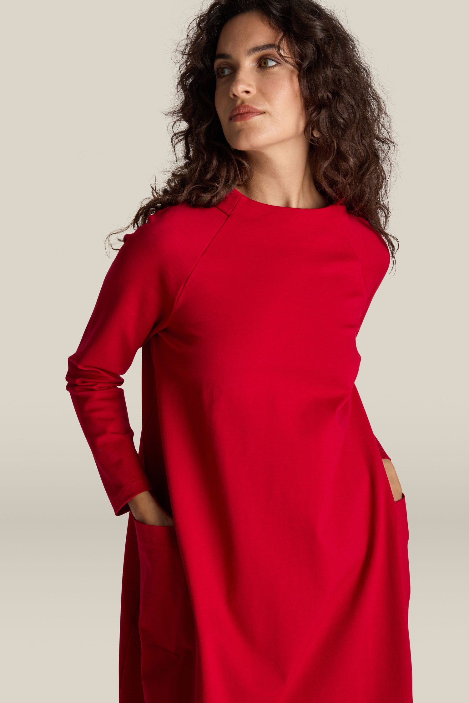 A person with curly hair stands and looks off-camera, wearing the Ponte Pocket Dress in a striking red color. The versatile silhouette of the Ponte Pocket Dress, enhanced by its practical pockets, adds to its effortless charm.