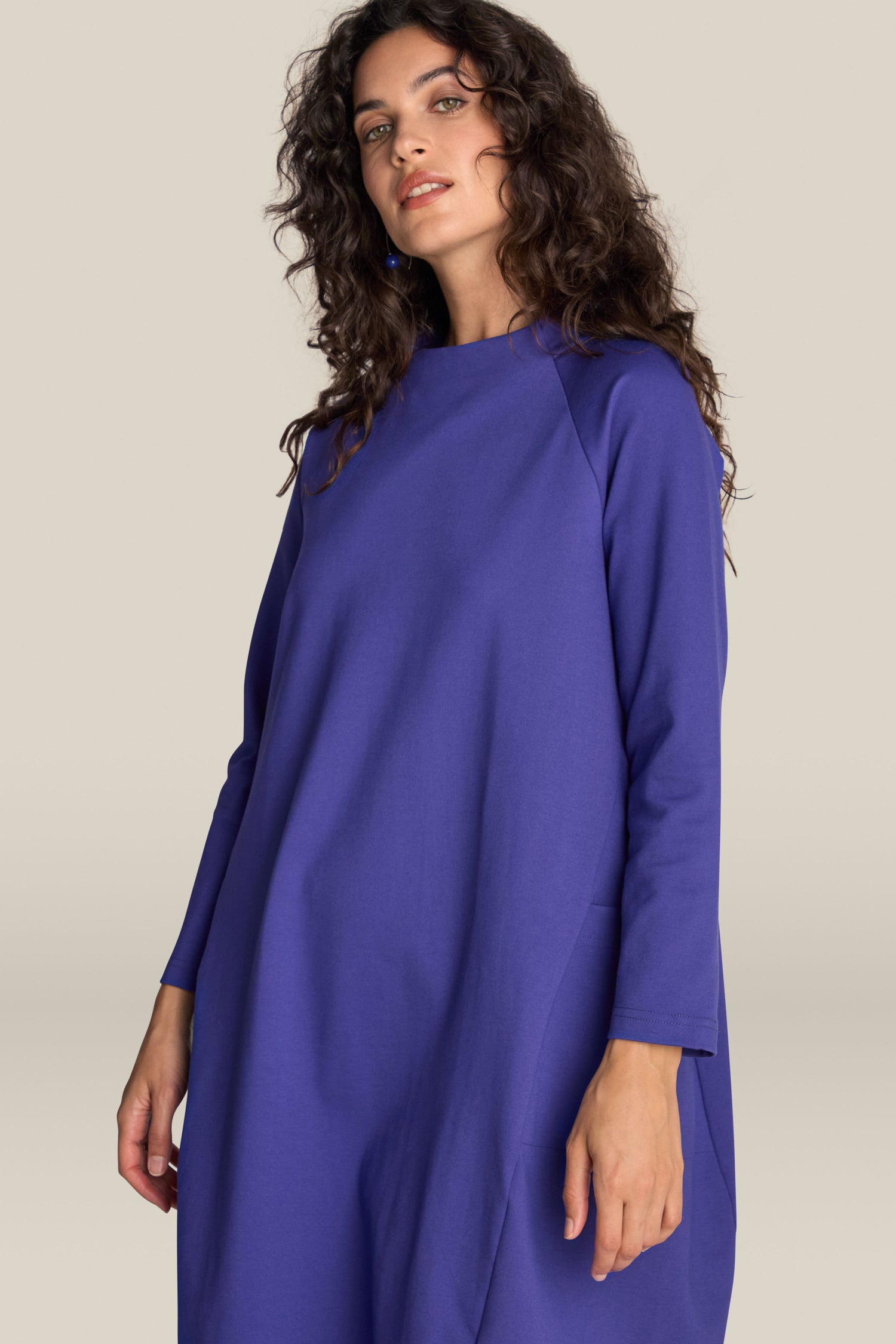 A person with long curly hair wears a loose, long-sleeved Ponte Pocket Dress in blue and stands against a plain background.