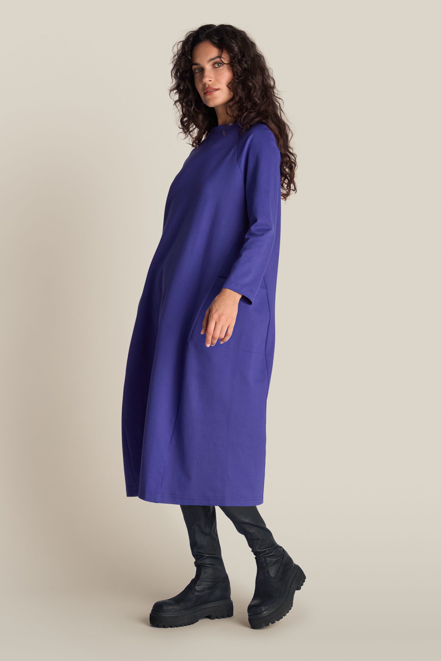 A person with long curly hair is wearing a Ponte Pocket Dress in purple, featuring a loose-fitting design with long sleeves and a cocoon shape. They are also wearing black knee-high boots, standing against a plain background.