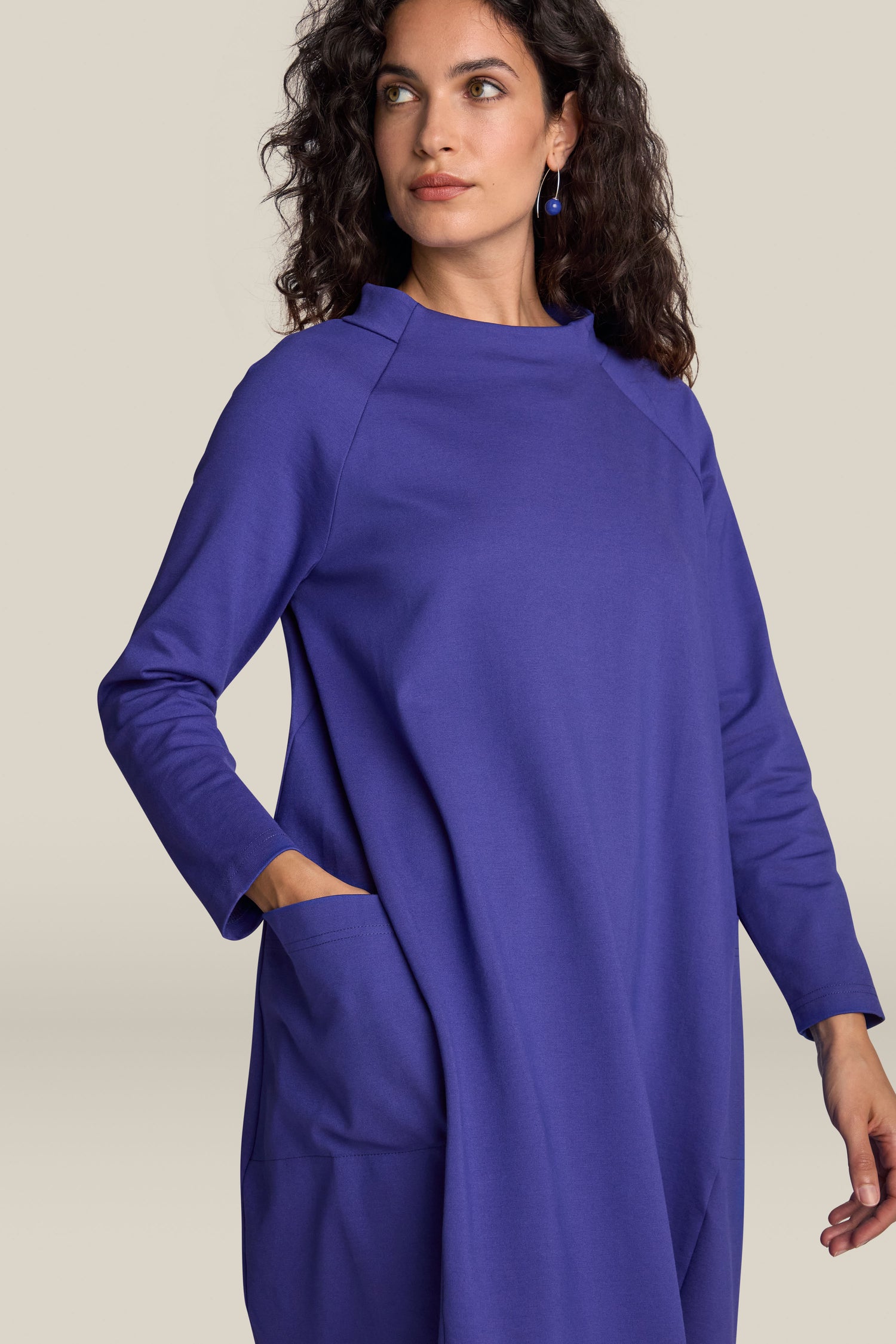 A person with curly hair, wearing the long-sleeved Ponte Pocket Dress in blue, featuring a stand-up collar and versatile silhouette, poses with one hand in their pocket while looking to the side.