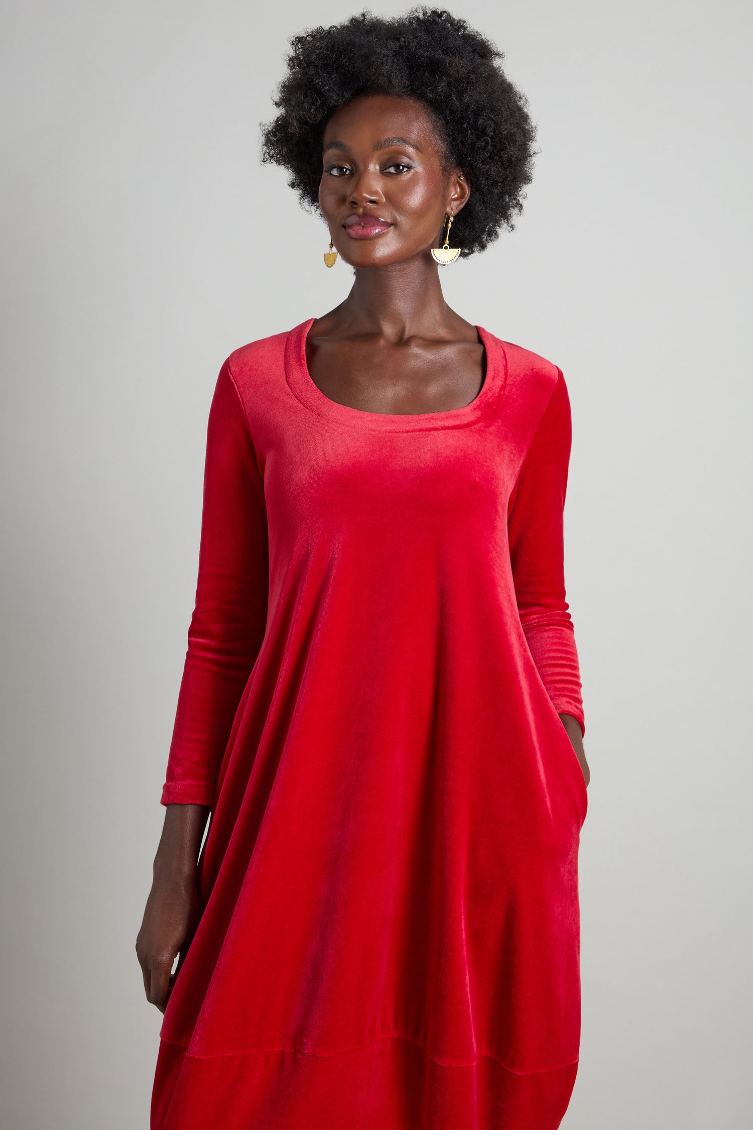 A person is dressed in a vibrant red Velvet Jersey Bubble Dress, complete with long sleeves and a chic horseshoe neckline. They pose confidently with their hands in the dress's pockets against a plain backdrop, highlighting the elegant flow and comfortable fit of its plush stretchy velvet fabric.