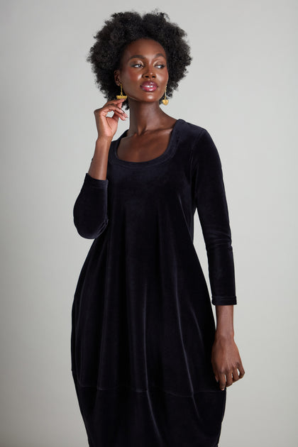 A person elegantly poses against a neutral backdrop, showcasing the luxurious Velvet Jersey Bubble Dress in black. With one hand near their face, they highlight the dress's sophisticated horseshoe neckline.