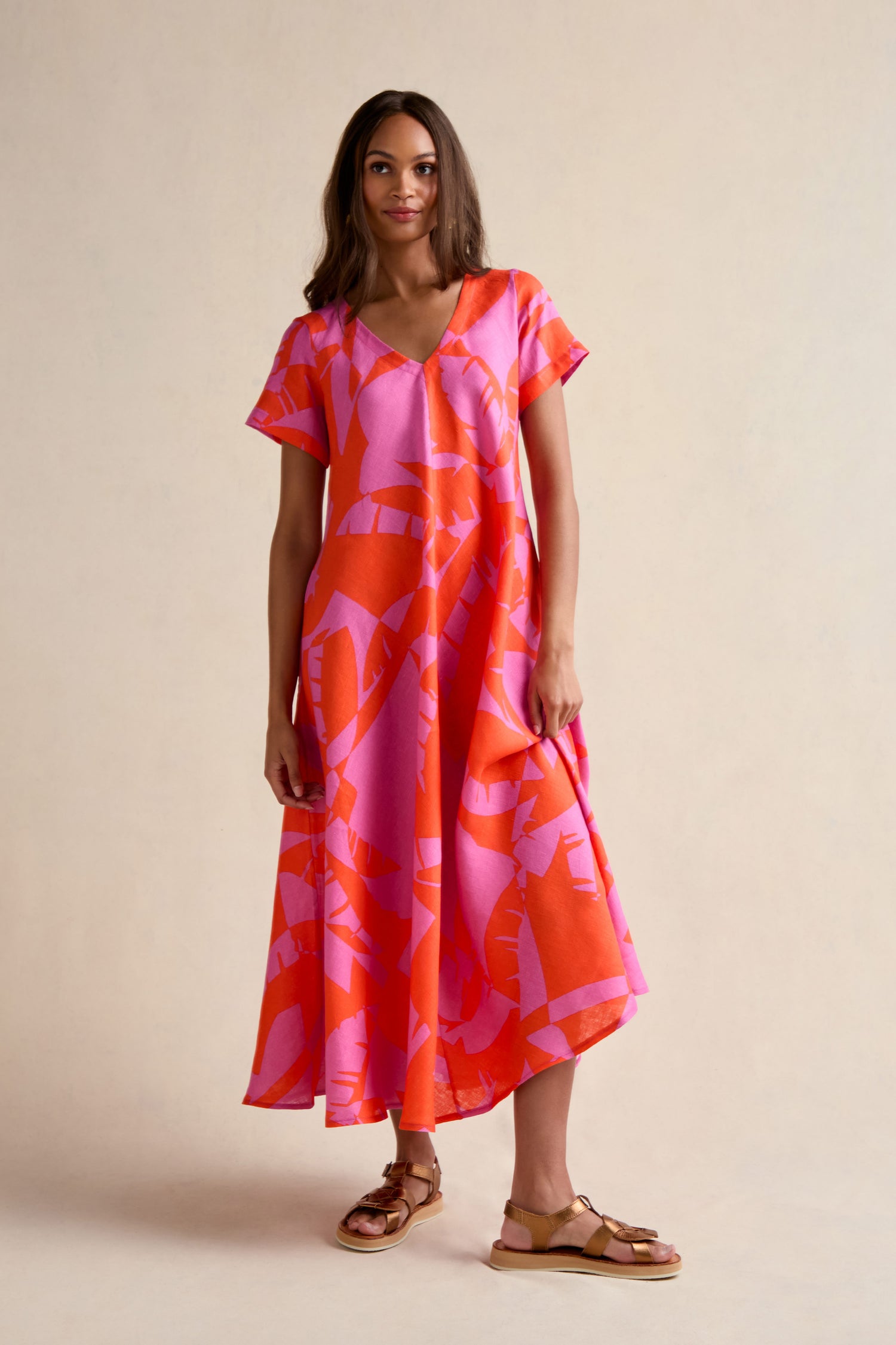 A woman stands in a studio wearing the Palm Mirage Linen Flared Dress, featuring a loose fit, short sleeves, and pink-orange palm print, paired with tan sandals.