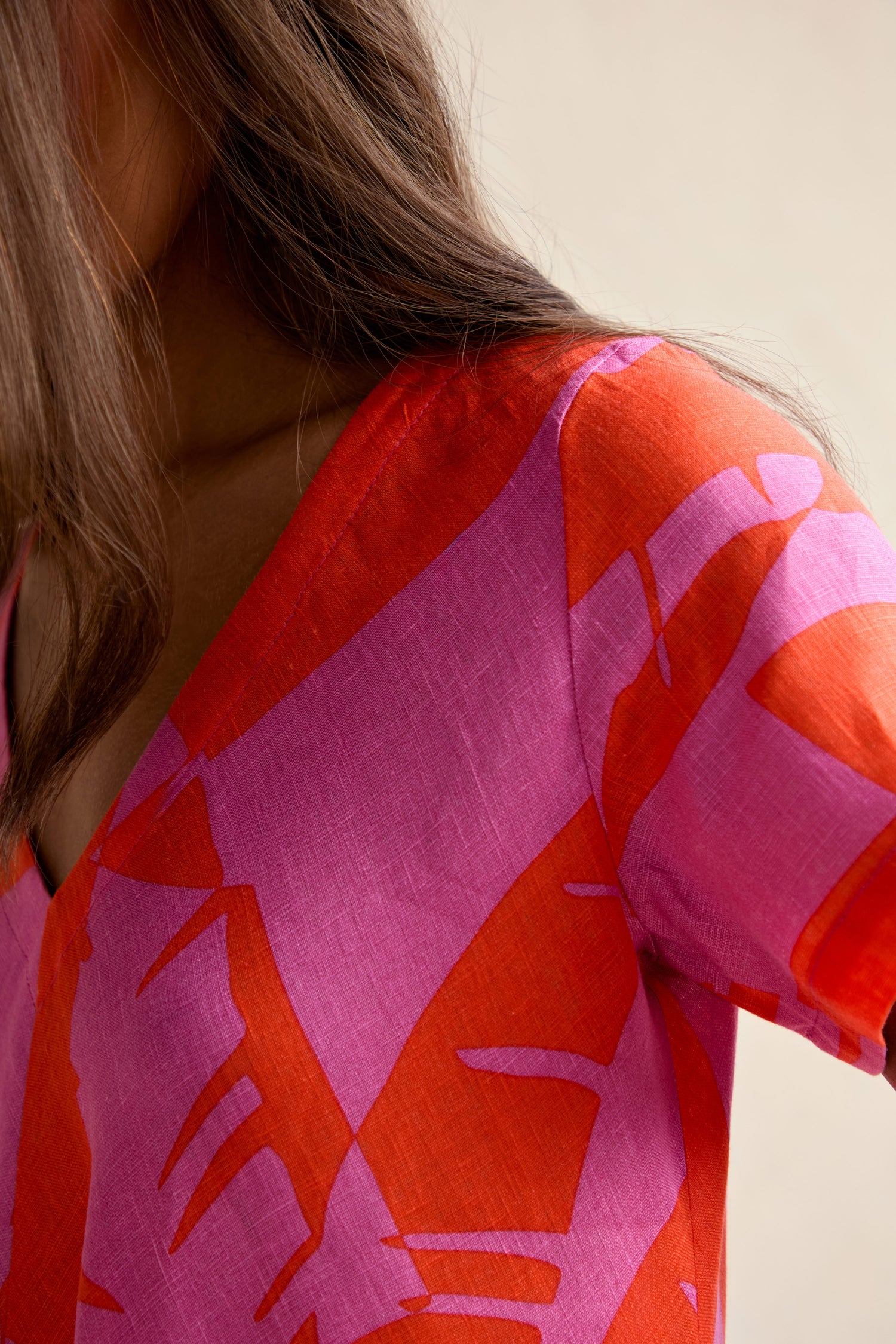 Person wearing the Palm Mirage Linen Flared Dress, a V-neck fit-and-flare garment with a bold pink and red abstract pattern; only the upper torso and part of the face are visible.