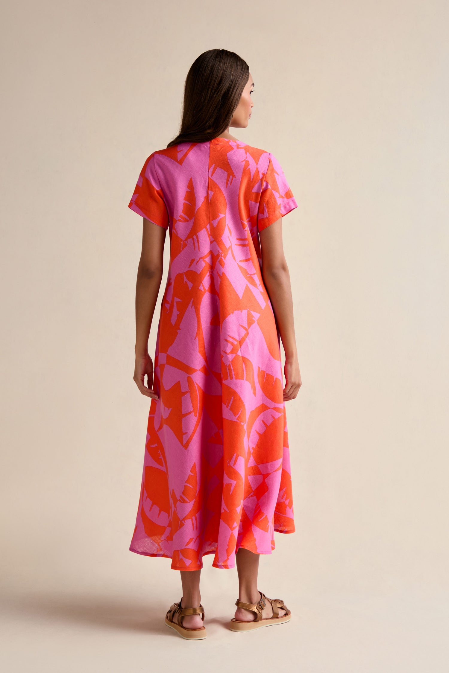 A woman stands with her back to the camera in the Palm Mirage Linen Flared Dress, featuring a pink and orange palm print and a fit-and-flare silhouette, paired with flat sandals against a neutral background.