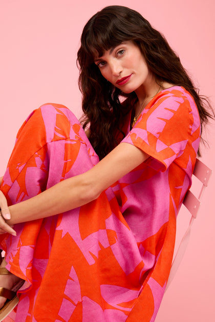 A woman sits on a pink chair against a pink background, wearing the Palm Mirage Linen Flared Dress featuring a loose fit, short sleeves, and a bold red and pink tropical abstract pattern.