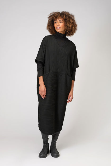 A person wears the Textured Wave Jersey Dress, a loose black kaftan-inspired dress in pleat-effect viscose jersey, over a black turtleneck and boots, standing against a plain light background.