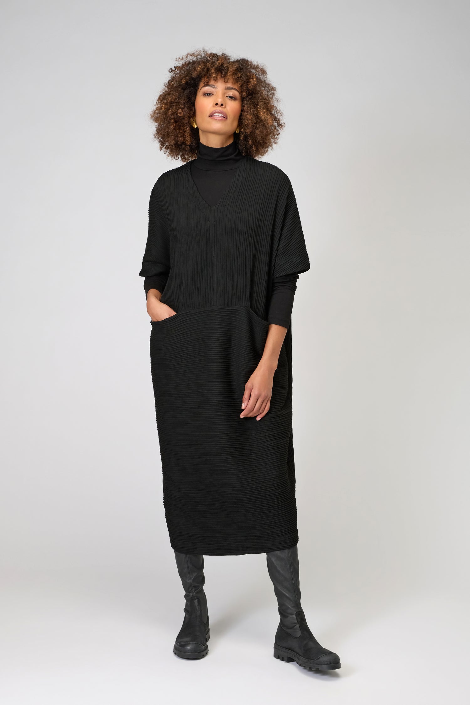 A woman stands against a light gray background, wearing the loose-fitting Textured Wave Jersey Dress over a black turtleneck and boots. One hand in her pocket highlights the dress’s relaxed, kaftan-inspired silhouette.