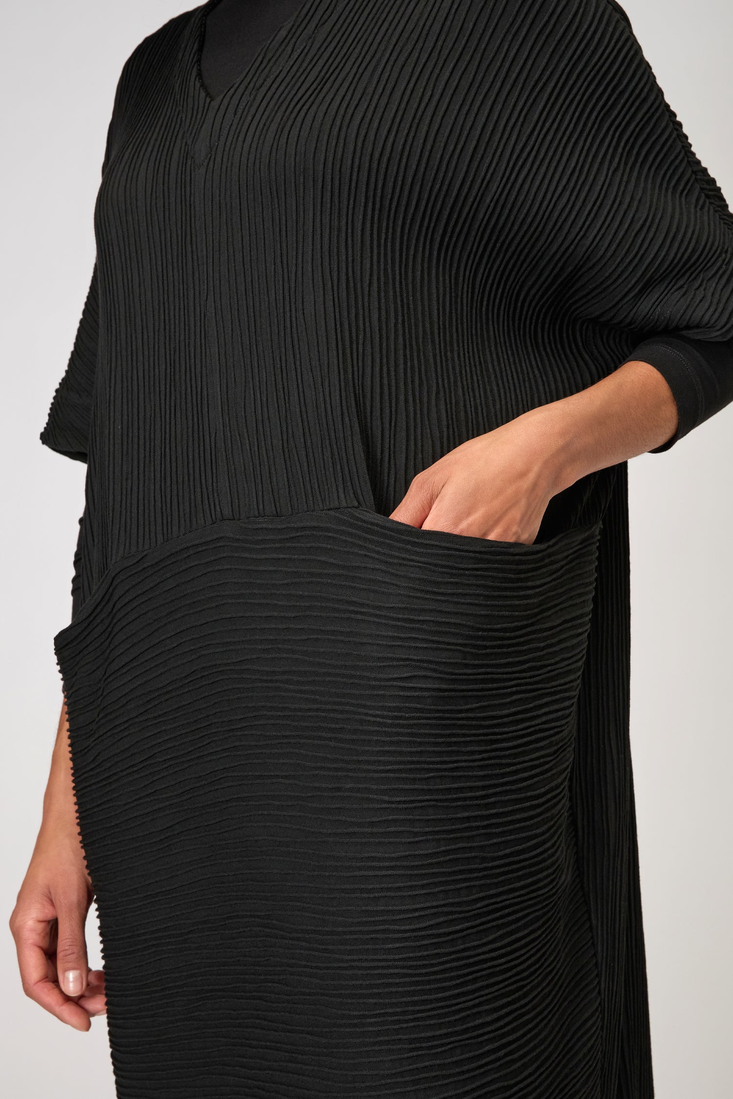 A person wearing the Textured Wave Jersey Dress, a black kaftan-inspired viscose piece featuring pleat effects and a large front pocket with one hand placed inside.