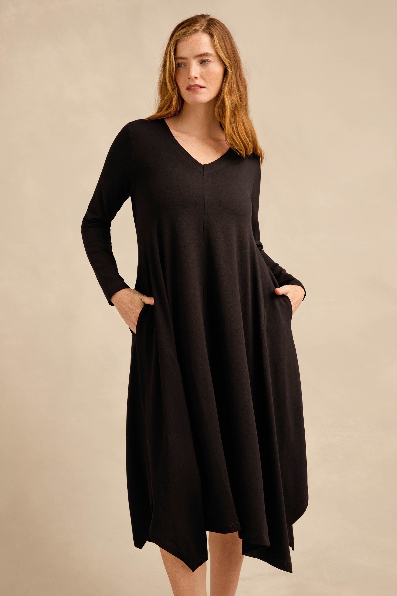 A woman wearing the Fluid Crepe Flared Dress—featuring long sleeves, a black crepe fabric, V-neckline, practical pockets, and a subtle handkerchief hem—stands against a plain beige background.