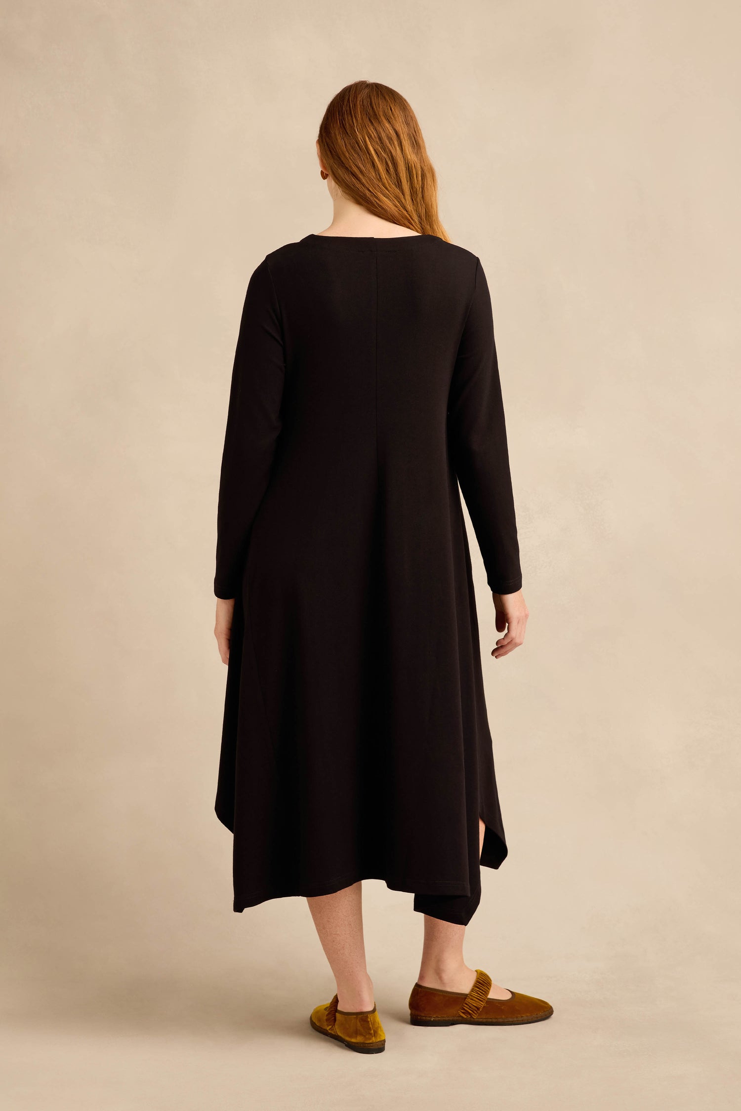 A person with long hair stands facing away, wearing the Fluid Crepe Flared Dress in black and brown shoes against a neutral background.