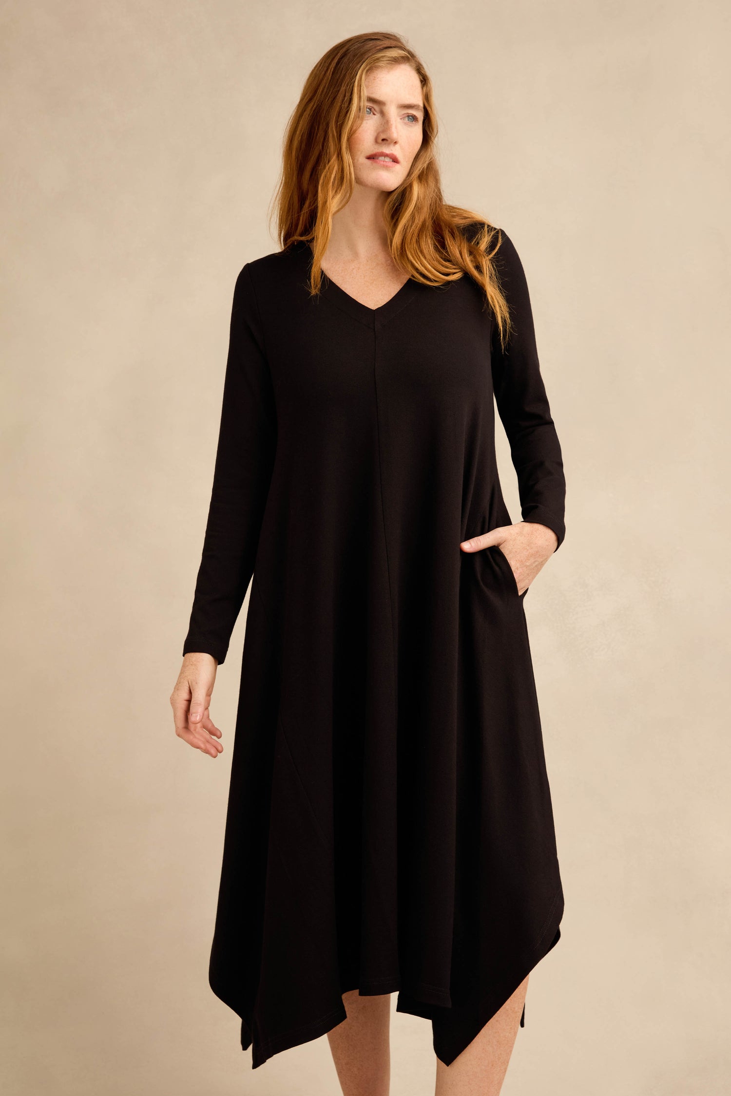 A woman stands against a plain beige background wearing the Fluid Crepe Flared Dress, a long-sleeved black crepe dress featuring a V-neckline and an ankle-length asymmetrical handkerchief hem.