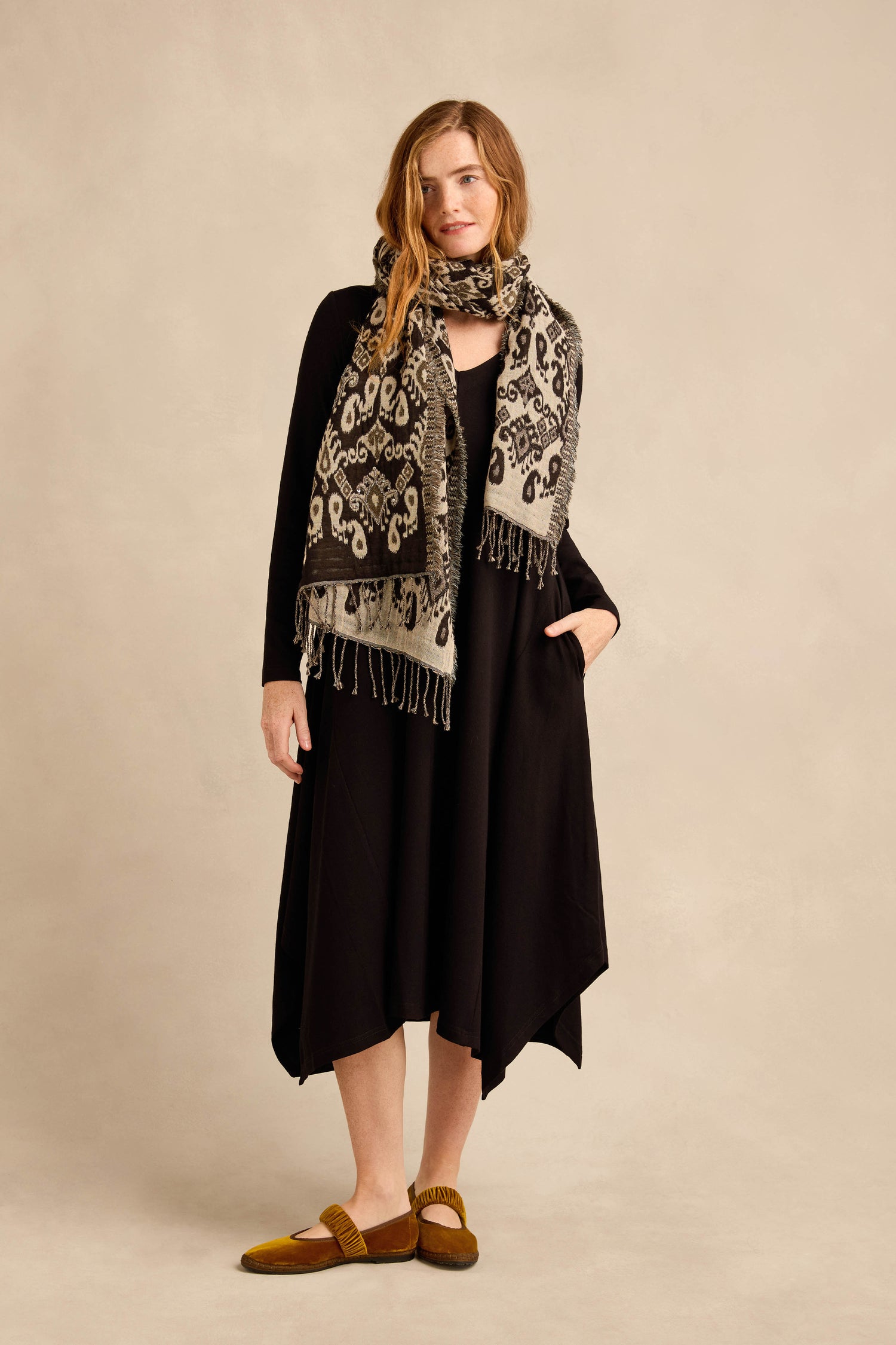 A woman stands against a beige background wearing the Fluid Crepe Flared Dress with a handkerchief hem, paired with a patterned scarf and brown shoes. She has one hand in her pocket and a relaxed expression.