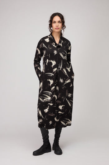 Woman wearing the Painted Brushstrokes Jersey Bubble Dress—black, long-sleeve, with white hand-painted brushstrokes and pockets—paired with black boots, stands against a plain light background.