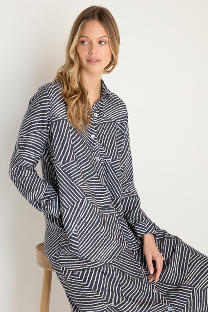A woman with long, wavy blonde hair sits on a wooden stool wearing the Linear Prism Shirt Dress—a long-sleeve, brushed viscose shirt dress made in England featuring bold black and white geometric stripes.