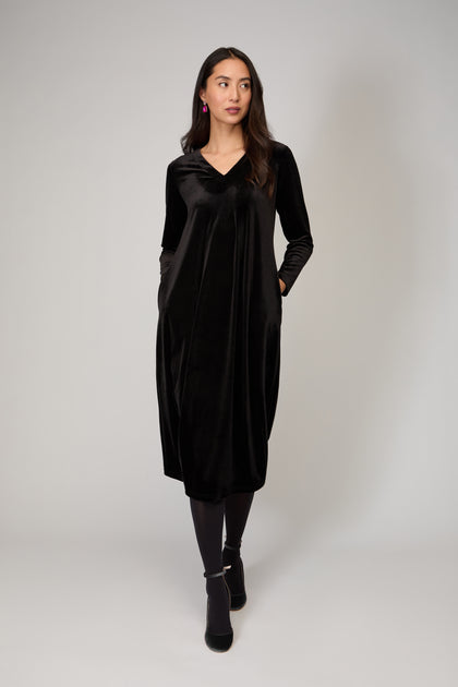 A woman wears the Velvet Jersey Bubble Dress—long-sleeved and black, with pockets—paired with black tights and shoes, standing against a plain light background for an effortless evening look.