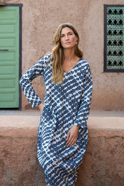 A woman with long wavy hair wears a blue and white Diamond Tie Dye Jersey Bubble Dress, standing against a textured beige wall with a green door and decorative window in the background.