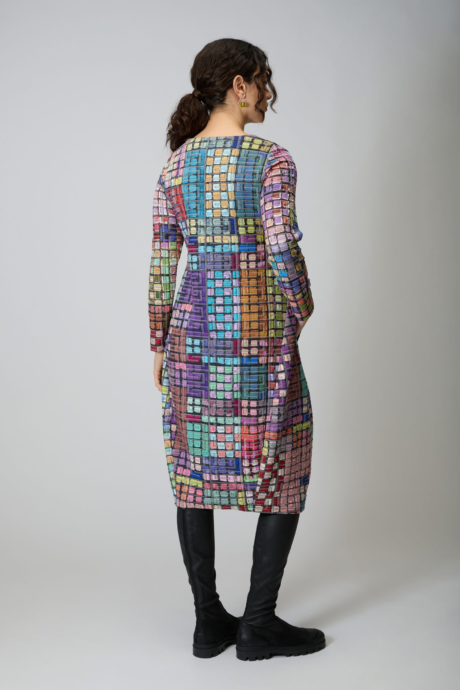 A person with curly hair in a ponytail wears the Coloured Glass Tiles Jersey Bubble Dress with long sleeves and a checkered pattern, paired with black knee-high boots, facing away from the camera against a plain background.