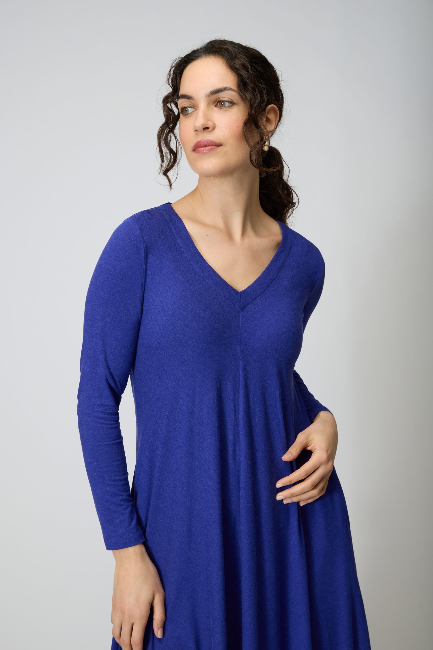A woman with curly dark hair models the Soft Viscose Jersey Bubble Dress in royal blue, featuring long sleeves and a V-neck, standing against a plain light background and gazing slightly to the side.