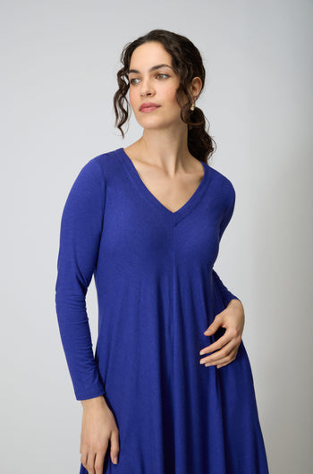 A woman with curly dark hair models the Soft Viscose Jersey Bubble Dress in royal blue, featuring long sleeves and a V-neck, standing against a plain light background and gazing slightly to the side.