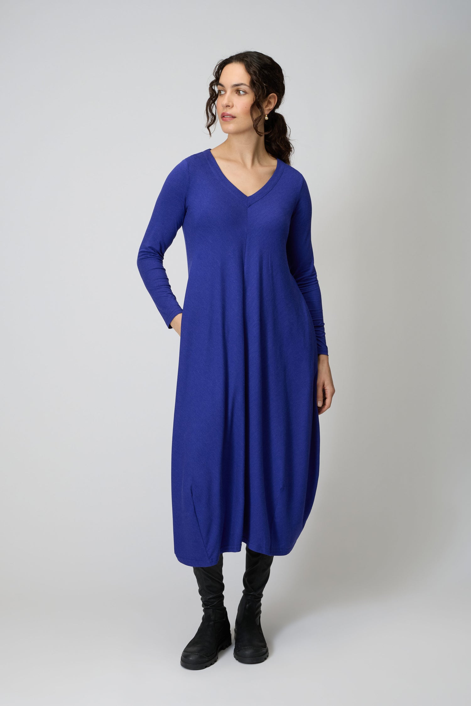 A woman stands against a plain background wearing the Soft Viscose Jersey Bubble Dress, featuring long sleeves, a V-neck, and a bubble silhouette in blue, paired with black boots.