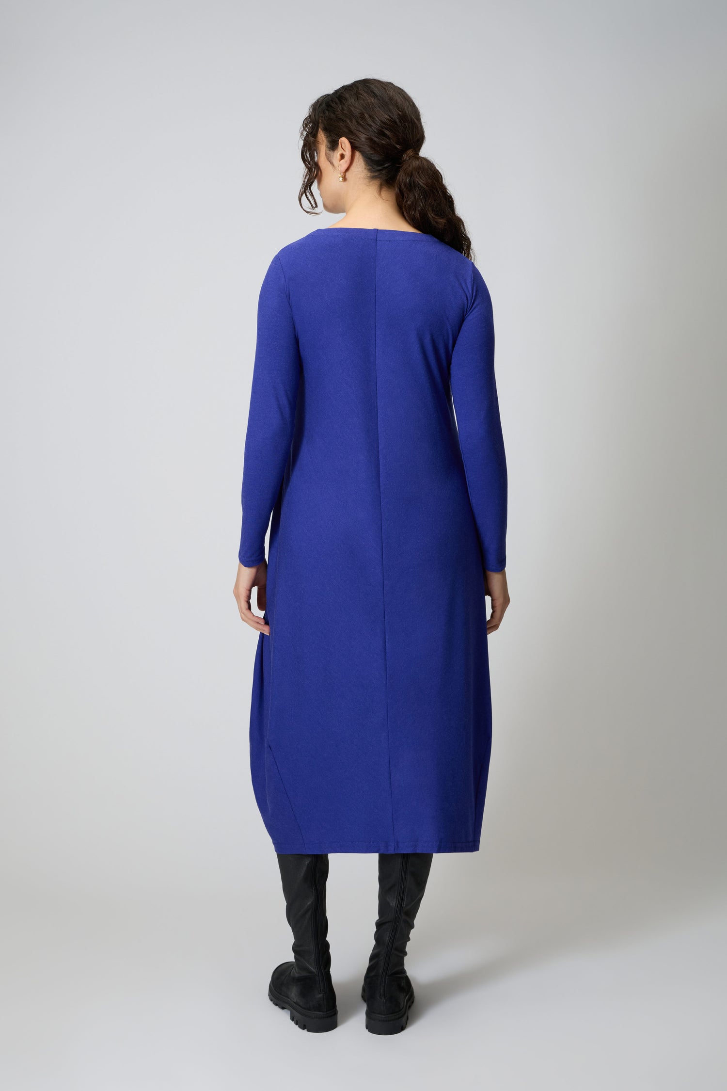 A woman faces away, wearing the Soft Viscose Jersey Bubble Dress—a luxurious, long-sleeved blue midi with a subtle bubble silhouette—paired with black knee-high boots against a plain backdrop.