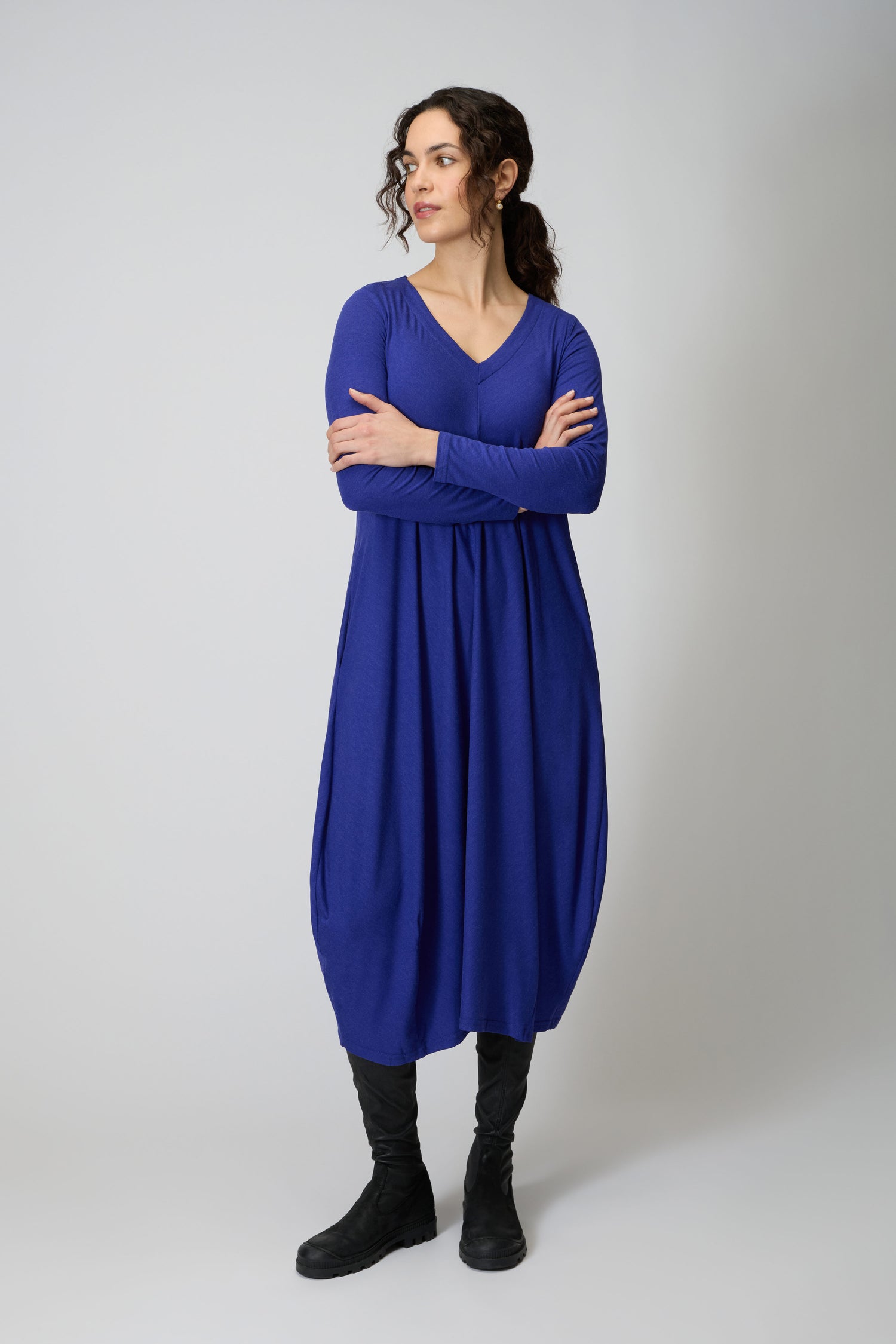 A woman stands with arms crossed, wearing the Soft Viscose Jersey Bubble Dress in blue and black boots, looking to her left against a plain light background.
